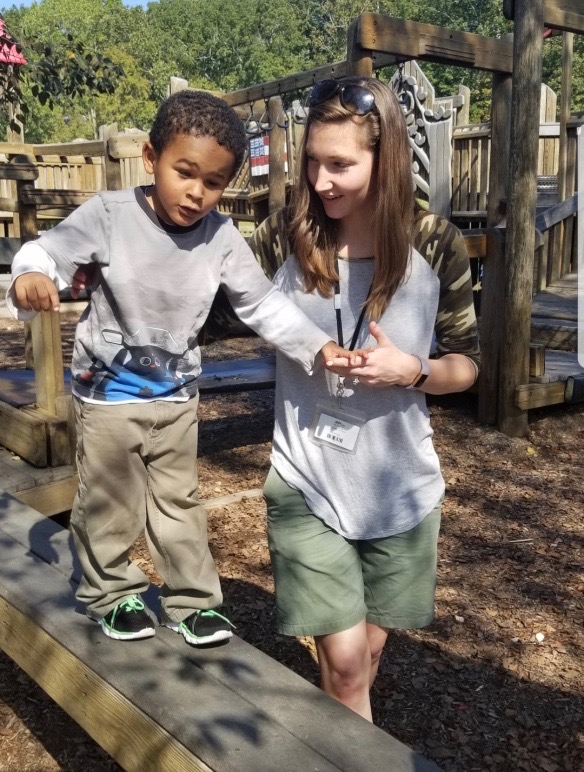 Occupational Therapist assists a young, male student across a balance beam at a park. 
